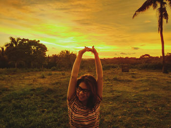 Portrait of woman with arms raised on field against sky during sunset
