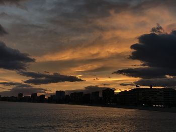 Sea by buildings against dramatic sky during sunset