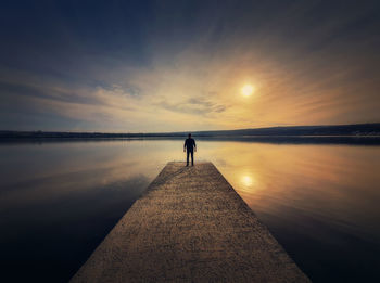 Man standing alone on the pier facing the sunset reflected in the calm lake water. scenic view