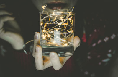 Midsection of woman holding glass with illuminated string light