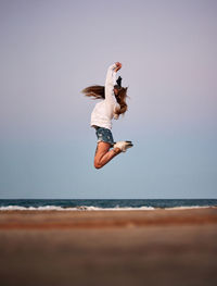 Ground level side view of carefree female traveler in moment of jumping above sandy shore on background of seascape in summer evening