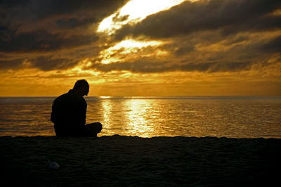 Silhouette man sitting on beach against sky during sunset