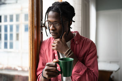 Thoughtful black man looks out window touches chin holding cup thinks about important work matters.