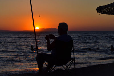 Silhouette fisherman sitting on beach against sky during sunset