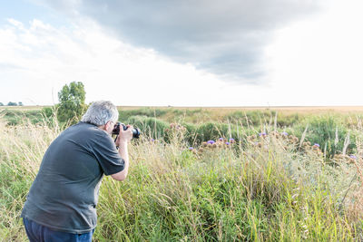 Rear view of a gray haired mature man taking camera shots of thistles in a field with a cloudy sky.