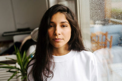 Young thoughtful ethnic female standing near window with raindrops and looking at camera