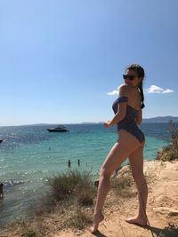 Side view of young woman in swimwear standing at beach against sky during sunny day