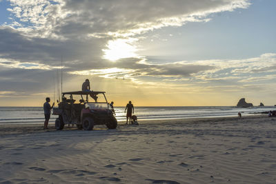 People by car at beach against sky during sunset