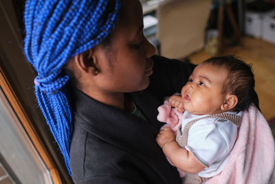 African american female with long blue hair standing at window and kissing newborn baby wrapped in soft towel