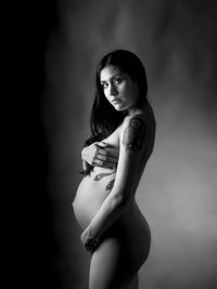 Side view portrait of naked pregnant young woman standing against black background