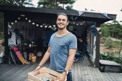 Portrait of happy young man carrying wooden crate on porch in back yard