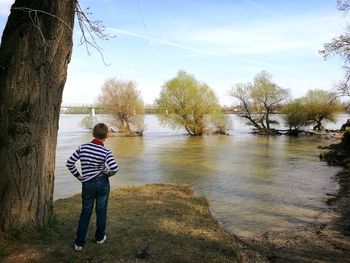 Rear view of boy standing by tree against river