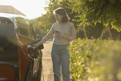 Woman with mobile phone charging electric car at roadside