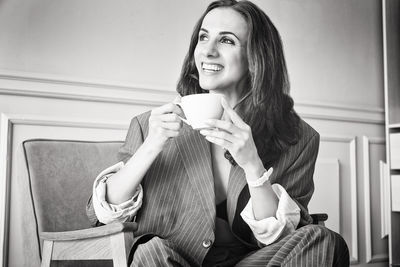 Ballerina in a gray striped suit with a cup of coffee smiling and dreaming is sitting in an armchair