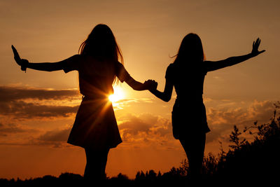 Silhouette friends holding hands standing on land against sky during sunset