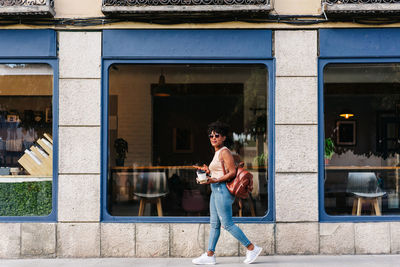 Full body of trendy young ethnic female millennial with dark afro hair in stylish outfit and backpack using mobile phone while walking on city street with cups of takeaway coffee