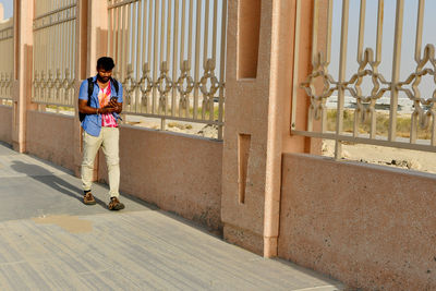 Young man using mobile phone while walking on footpath during sunny day