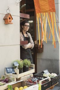 Female owner with arms crossed standing at entrance of store by vegetable crates