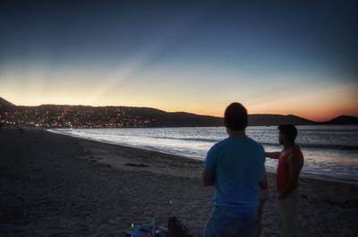 Rear view of friends standing at beach against sky during sunset