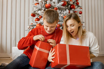 Young couple sitting near christmas tree and starting unpack presents together with excitement