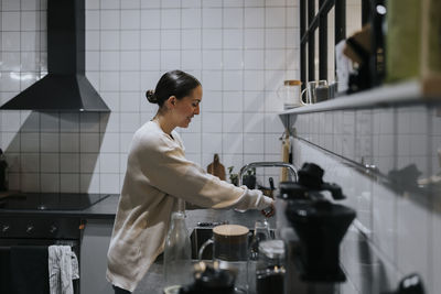 Side view of smiling businesswoman washing glass at sink in office kitchen