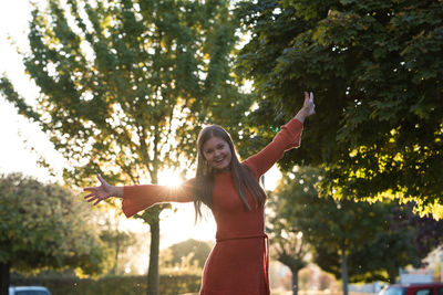 Portrait of smiling woman with arms outstretched standing against trees