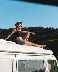 Full body side view of of pleasant barefoot thoughtful hippie female traveler sitting with eyes closed on roof of white camping van parked in nature during road trip