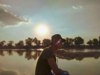Side view of young man sitting in lake against sky during sunset
