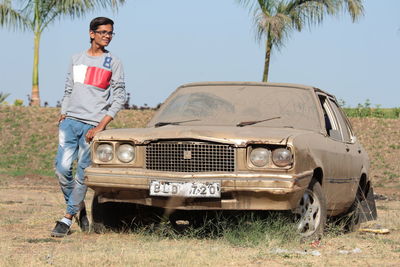 Smiling young man standing by damaged car on field