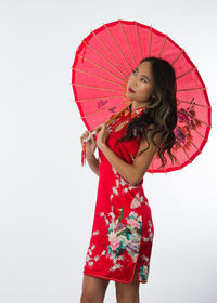 Thoughtful young woman in red dress holding traditional umbrella against while background