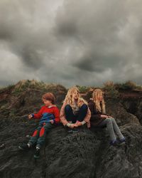 Low angle view of friends sitting on rock against cloudy sky