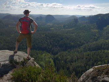 Travelling man wearing brown hat, green t-shirt, shorts and sandals with red backpack stay on cliff