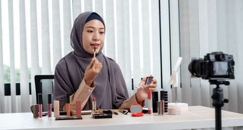 Young woman blogging while applying lipstick at office