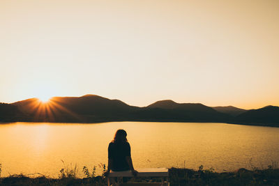 Rear view of woman sitting by lake against sky during sunset