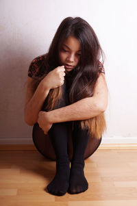 Full length of sad woman sitting on hardwood floor against wall at home