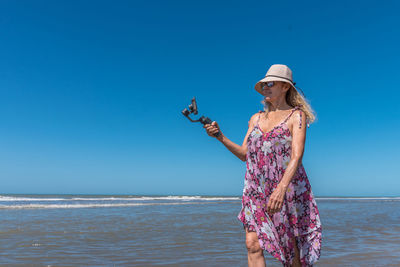 Blond woman in hat and summer dress taking a picture while strolling on the beach