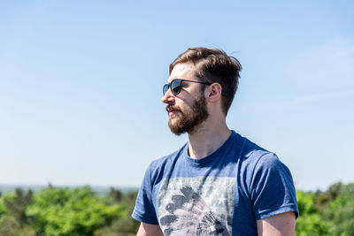 Bearded young man looking away while standing against sky during sunny day