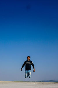 Full length of young man jumping at sandy beach against clear blue sky