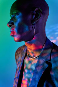 Side view of serious bald african american slim male in necklace and stylish outfit standing with eyes closed in modern studio with dim illumination and glowing lights on green and blue background