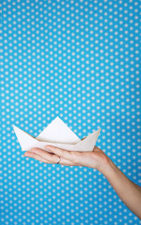 Cropped hand of woman holding paper boat against blue wall