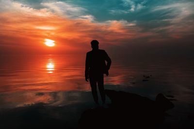 Silhouette man standing at beach against dramatic sky during sunset