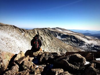 Rear view of backpacker looking at mountains while sitting on rocks against blue sky