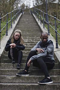 Full length of multi-ethnic couple checking wristwatch and mobile phone while sitting on steps