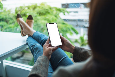 Mockup image of a woman holding mobile phone with blank white desktop screen while sitting in office