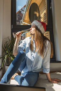 A girl sits in a room on the floor in front of a laptop in a santa hat and a glass of champagne 