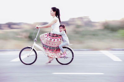 Daughter with mother riding bicycle on street against field