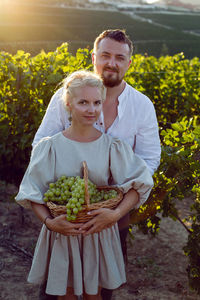 Man and a woman in white clothes stand in a grape field at sunset holding a basket of grapes