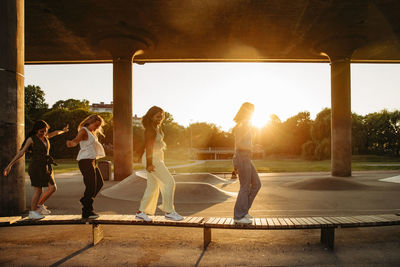 Full length of teenage girls walking on bench under bridge during sunset