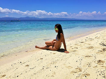 Side view of woman wearing bikini sitting at beach against sky