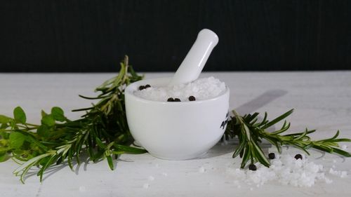 Close-up of rock salt with peppercorn in mortar and pestle by rosemary on table against black background